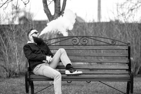 Vape. Young brutal man with large beard and fashionable haircut in sunglasses smokes an electronic...