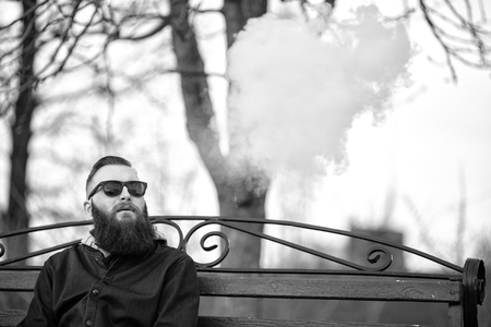 Vape. Young brutal man with large beard and fashionable haircut in sunglasses smokes an electronic cigarette on the red bench in the city park. Steam cloud. Lifestyle. Black and white photo.の写真素材