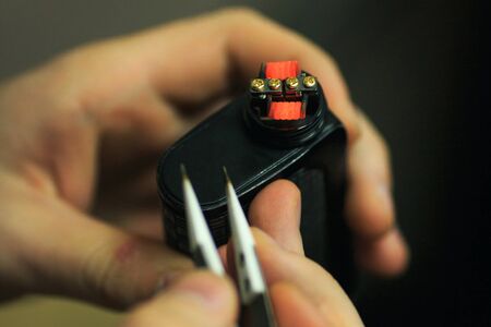 Vape after-sales service of the e-cigarette. Male hands regulate electronic cigarette coils with pincette. Glowing spiral for background. Personal vaporizer. Close up.の写真素材