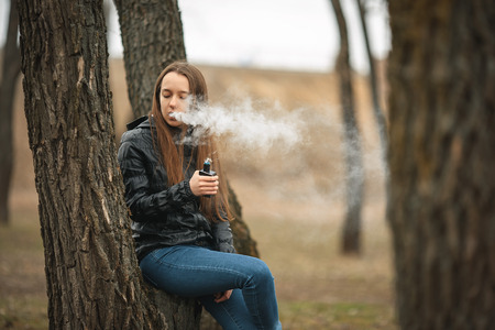 Vape. A young beautiful white girl smokes an electronic cigarette near a tree. Lifestyle.