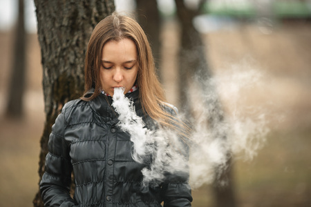 Vape. A young beautiful white girl smokes an electronic cigarette near a tree. Lifestyle.の写真素材