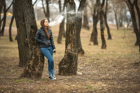 Vape. A young beautiful white girl smokes an electronic cigarette near a tree. Lifestyle.
