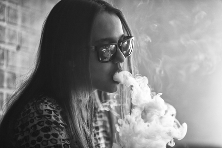 Vape. Young handsome white girl in sunglasses is admiting puffs of steam from the electronic cigarette. Vaping. Teenager. Black and white photo.の写真素材