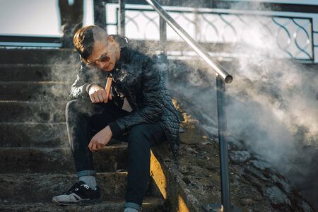 Vape. A young handsome guy sits on the stairs and blows steam from an electronic cigarette. Lifestyle.の写真素材