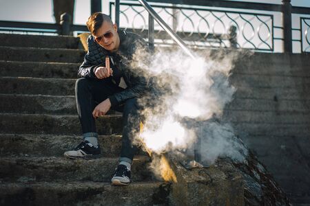Vape. A young handsome guy sits on the stairs and blows steam from an electronic cigarette. Lifestyle.の写真素材