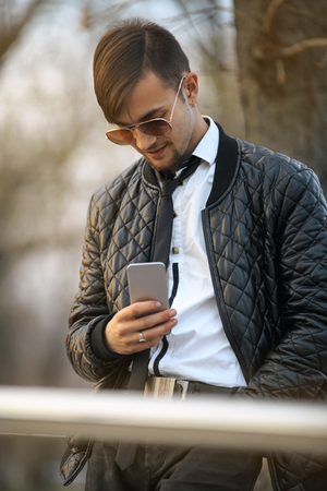 Gadget man with mobile phone. Portret of a handsome young white guy with modern haircut in aviator sunglasses posing with smartphone at sunset. Lifestyle.の写真素材