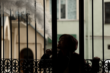 Vape man. Silhouette of a young white guy in the hood vaping and letting off steam from an electronic cigarette near a vintage metal gate. Lifestyle.の写真素材