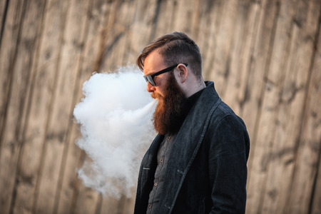 Vape man. Outdoor portrait of a young brutal white guy with large beard and in sunglasses letting puffs out of steam from an electronic cigarette opposite the old wooden fence in the village. Vaping.の写真素材