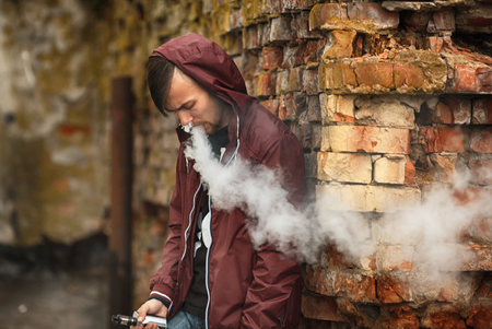 Vape man. Photo of a handsome young white guy in the hood vaping and letting off steam from an electronic cigarette near old destroyed red brick wall. Lifestyle.の写真素材
