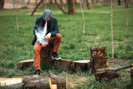 Vape man. Photo of a brutal bearded young man having rest after chop firewood and vaping an...