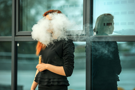Vape teenager. Young pretty white girl with red curly hair and modern haircut in black dress vaping an electronic cigarette opposite city background.の写真素材