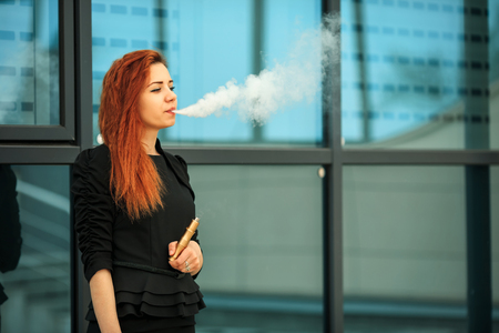 Vape teenager. Young pretty white girl with red curly hair and modern haircut in black dress vaping...