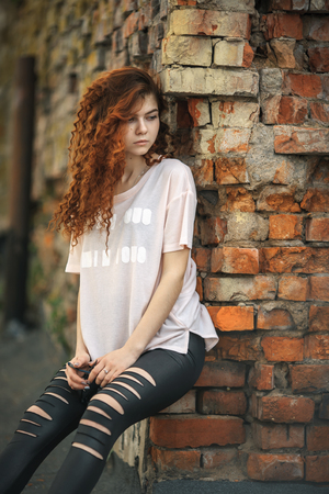 Beautiful teenager. Outdoor portrait of a pretty young white girl with red curly hair posing near old destroyed red brick wall. Lifestyle.の写真素材