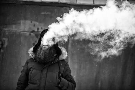 Vape bearded man in real life. Portrait of young guy with large beard in glasses and a cap vaping an electronic cigarette and letting out steam opposite the garage in the winter. Black and white.の写真素材
