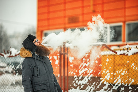 Vape bearded man in real life. Portrait of young guy with large beard in glasses and a cap vaping an...