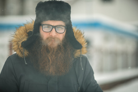 Vape bearded man in real life. Portrait of young guy with large beard in glasses and a black cap vaping an electronic cigarette and letting out steam in the winter.の写真素材