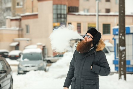 Vape bearded man in real life. Portrait of young guy with large beard in glasses and a black cap vaping an electronic cigarette and letting out steam in the winter.の写真素材