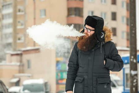 Vape bearded man in real life. Portrait of young guy with large beard in glasses and a black cap vaping an electronic cigarette and letting out steam in the winter.の写真素材
