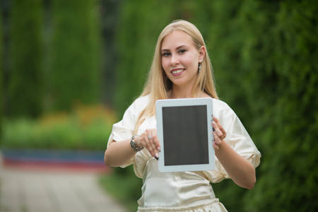 Young pretty blonde girl in a dress with a tablet in her hands between thuja on the street in summer.の写真素材