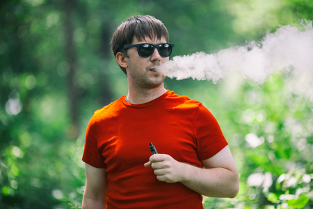 A young white bearded man in red t-shirt and in sunglasses is vaping an electronic cigarette in the forest in the summer. Close up.の写真素材