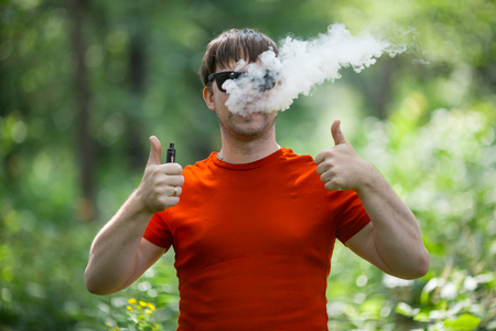 A young white bearded man in red t-shirt and in sunglasses is vaping an electronic cigarette in the forest in the summer. Close up.の写真素材