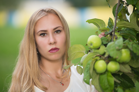 Portrait of a young cute blonde white girl near the tree with green apples in the garden in summer.の写真素材