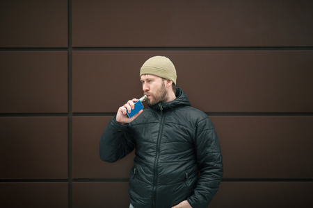 Vape man. Young bearded white guy in the cap and black jacket is smoking an electronic cigarette...