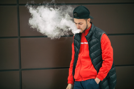Vape teenager. Young handsome bearded white man in black cap smoking an electronic cigarette and letting off the steam opposite the modern brown background in autumn.の写真素材