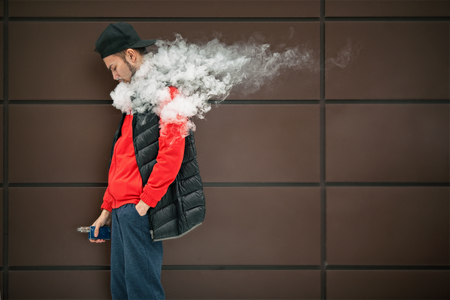 Vape teenager. Young handsome bearded white man in black cap smoking an electronic cigarette and letting off the steam opposite the modern brown background in autumn.の写真素材