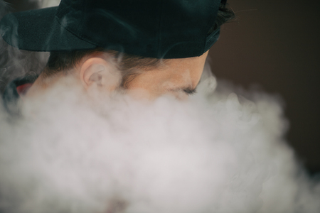 Vape teenager. Young white man in black cap smoking an electronic cigarette and letting off the...