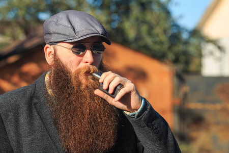 Vape bearded man in real life. Portrait of young guy with large beard in sunglasses and a gray cap vaping an electronic cigarette on the street in the village in the autumn.の写真素材
