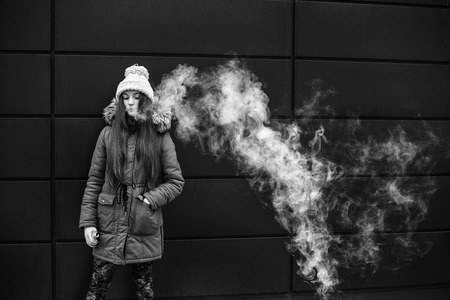 Vape teenager. Young pretty white girl smoking an electronic cigarette opposite modern background on the street in the winter. Badの写真素材