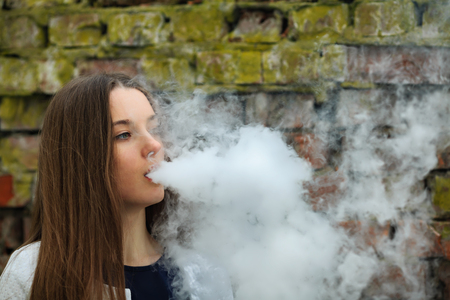 Vape teenager. Young pretty white girl in white cardigan smoking an electronic cigarette opposite destroyed brick wall on the street in the spring. Bad habit.の写真素材