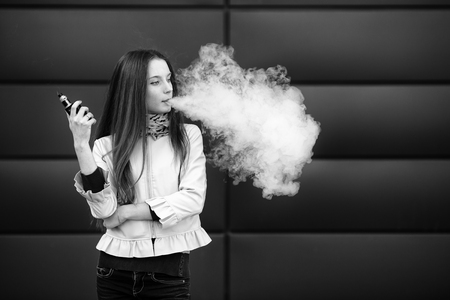 Vape teenager. Young pretty white girl in casual clothing smoking an electronic cigarette opposite modern background on the street in the spring. Bad habit. Vaping activity. Black and white.の写真素材