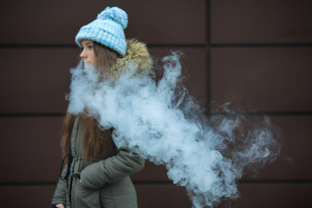 Vape teenager. Young pretty white girl smoking an electronic cigarette opposite modern brown background on the street in the winter. Close up.の写真素材