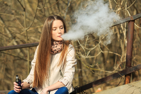 Vape teenager. Young pretty white girl in casual clothing smoking an electronic cigarette on the old wooden stairs on the street in the spring. Bad habit. Vaping activity.の写真素材