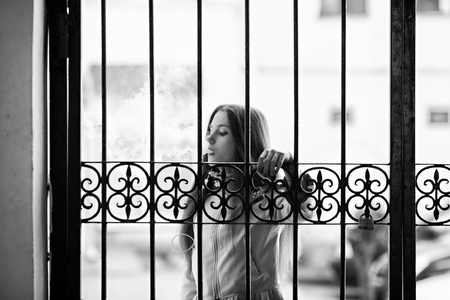Vape teenager. Young pretty white girl in casual clothing smoking an electronic cigarette near vintage metal gates on the street in the spring. Bad habit. Vaping activity. Black and white.の写真素材
