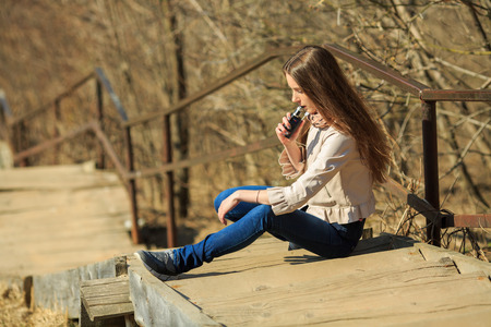 Vape teenager. Young pretty white girl in casual clothing smoking an electronic cigarette on the old wooden stairs on the street in the spring. Bad habit. Vaping activity.の写真素材