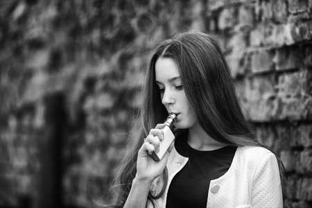 Vape teenager. Young pretty white girl in white cardigan smoking an electronic cigarette opposite brick wall on the street in the spring. Bad habit.  Black and white.の写真素材