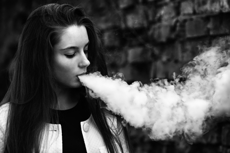 Vape teenager. Young pretty white girl in white cardigan smoking an electronic cigarette opposite brick wall on the street in the spring. Bad habit.  Black and white.の写真素材