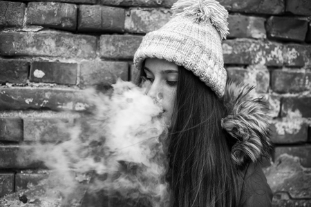 Vape teenager. Young pretty white girl in a cap and jacket smoking an electronic cigarette opposite brick wall on the street in the spring. Bad habit. Black and white.の写真素材