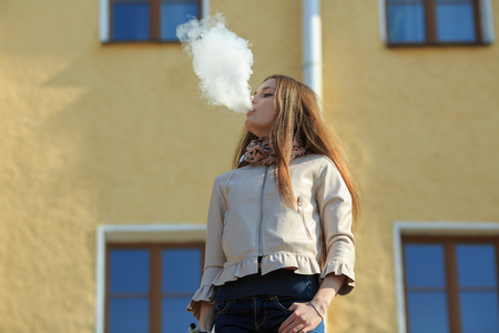 Vape teenager. Young pretty white girl in casual clothing smoking an electronic cigarette in front of a vintage building on a sunny day in the spring. Bad habit. Vaping activity.の写真素材