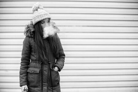 Vape teenager. Young pretty white girl in a cap and jacket smoking an electronic cigarette opposite modern background on the street in the winter. Bad habit. Black and white.の写真素材