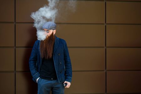 Vape bearded man. An adult man with a very long beard in a cap and sunglasses smokes an electronic...