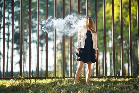 Vape teenager. Young cute girl in casual clothes smokes an electronic cigarette in front of a metal fence outdoors in the forest at sunset in summer. Bad habit. Stop vaping.の写真素材