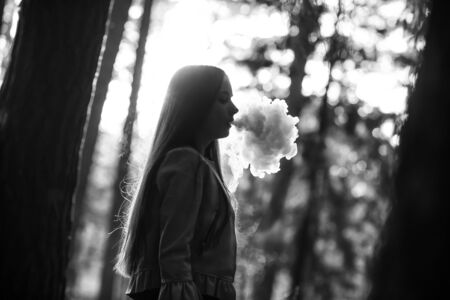 Vape teenager. Young cute girl in casual clothes smokes an electronic cigarette outdoors in the forest at sunset in summer. Bad habit that is harmful to health. Vaping activity. Black and white.の写真素材