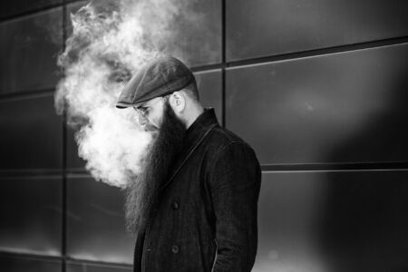 Vape bearded man. An adult man with a very long beard in a cap and sunglasses smokes an electronic cigarette on a sunny day outside. Bad habit that is harmful to health. Black and white.の写真素材