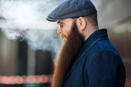 Vape bearded man. An adult man with a very long beard in a cap and sunglasses smokes an electronic cigarette on a sunny day outside. Bad habit that is harmful to health.の写真素材