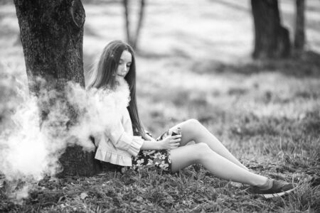 Vape teenager. Young cute girl in a dress sits near a tree and smokes an electronic cigarette outdoors in a park in spring. Bad habit that is harmful to health. Vaping activity. Black and white.の写真素材