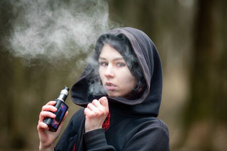 Vape teenager. Young pretty caucasian brunette girl smoking an electronic cigarette on the street in the spring. Deadly bad habit.の写真素材